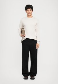 Hellgrauer Strickpullover, schwarze weite Hose, hält eine beigefarbene Tasche. Die Schuhe sind dunkelbraun, haben eine glatte Oberfläche und ein minimalistisches Design.