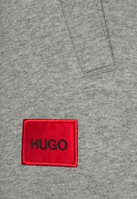 La tela del sudadera gris presenta un parche rectangular rojo con "HUGO" en letras negras. La textura es suave y con un ligero aspecto jaspeado.