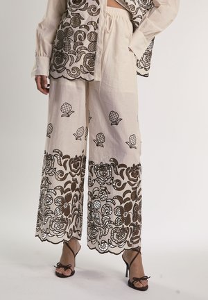 Beżowe spodnie o szerokich nogawkach z ciemnobrązową kwiatową haftem na dole oraz rozsianymi mniejszymi motywami kwiatowymi, w połączeniu z czarnymi sandałami na obcasie.