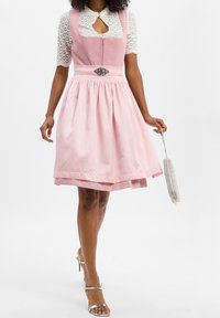 Femme portant une robe dirndl rose avec un chemisier en dentelle blanche, tenant une petite pochette perlée, et des sandales argentées à talons, marchant vers l'avant.