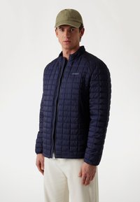 Jachetă quilted de culoare bleumarin cu guler înalt și fermoar frontal, având logo-ul brandului, asociată cu o făcută verde deschis și pantaloni albi.