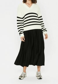 Stickad tröja med vita och svarta ränder, V-ringning, i kombination med en svart plisserad kjol och grå sneakers. Strukturerad tyg och avslappnad passform.