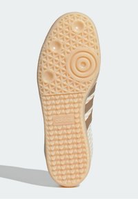 Semelle de chaussure Adidas en beige, présentant une adhérence en caoutchouc texturé avec des motifs circulaires et en losange, et un logo en relief au centre.