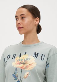 Svetle zelená mikina s květinovou grafikou, oversized střih, kulatý výstřih a nápis "SOW LOVE." Květinový design obsahuje jemné růžové a modré barvy.
