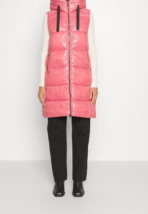 Kobieta ubrana w różową, błyszczącą pikowaną kamizelkę bez rękawów z kapturem, białą bluzkę z długim rękawem, czarne spodnie i czarne botki, stojąca na tle białej ściany.