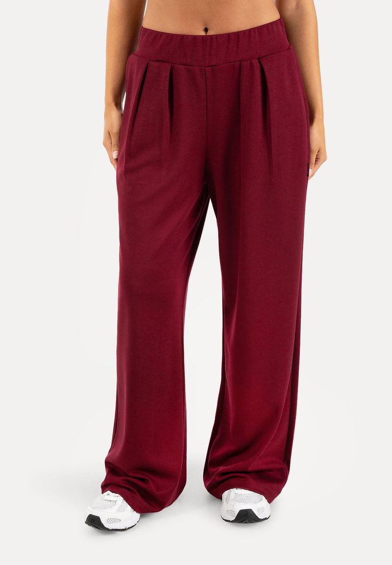 Partie inférieure du corps d'une personne portant un pantalon bordeaux ample à taille haute et des baskets blanches, debout sur un fond clair uni.