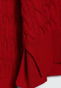 Pull en tricot rouge avec un motif en cable et des poignets côtelés. Le matériau est doux et texturé, avec une coupe ample pour le confort.