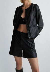 Czarna kurtka z ekologicznej skóry z plecionymi akcentami, z przodu zapinana na guziki, zestawiona z dopasowanymi czarnymi skórzanymi szortami.