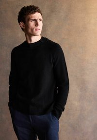 Schwarzer gestrickter Pullover mit geripptem Ausschnitt und Bündchen, über blauen strukturierten Hosen getragen, vor einem neutralen Hintergrund.