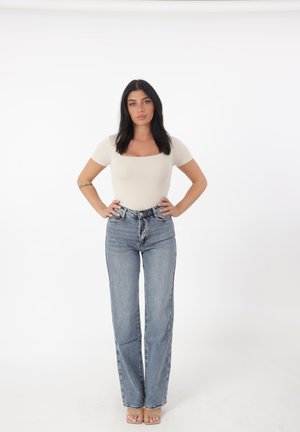 Top ajustado color beige claro con escote cuadrado, combinado con vaqueros de pierna recta de talle alto en azul. Sandalias transparentes completan el conjunto.