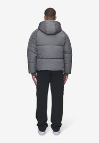 Uomo di spalle che indossa un giubbotto puffy grigio con cappuccio, pantaloni cargo neri e sneakers bianche su uno sfondo bianco uniforme.
