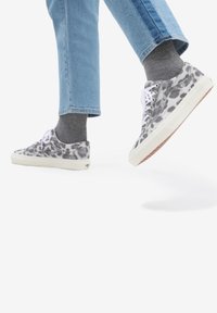 Gråprickiga sneakers med vita snören och gummisulor, kombinerade med ljusblå jeans och grå strumpor, i ett steg mot en vit bakgrund.