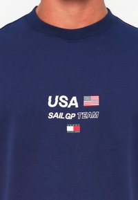 Navy blue t-shirt made of cotton with a crew neck. Features "USA" and "SAILGP TEAM" in white text, a flag graphic, and a small logo.