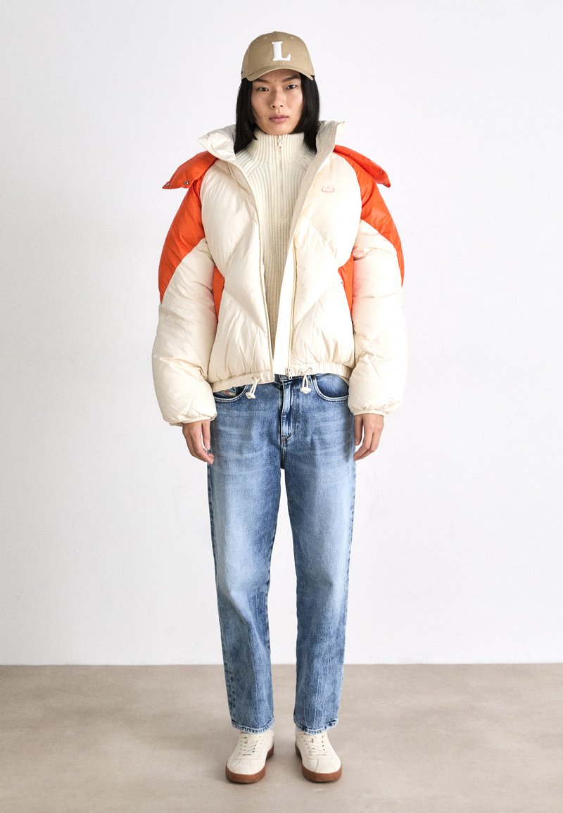 Giubbotto imbottito con sezioni bianche e arancioni, maniche ampie e gonfie. Indossato con un maglione crema, jeans azzurri chiari e sneakers crema. Suole marroni.