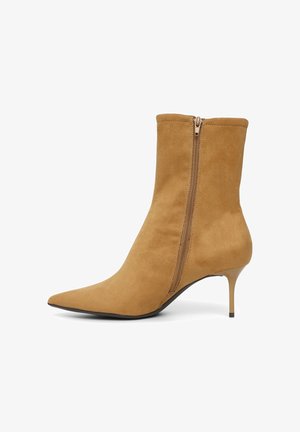 Bottine en suède beige avec un bout pointu, fermeture éclair sur le côté, et un talon fin. La texture lisse et la coupe ajustée accentuent son apparence élégante.