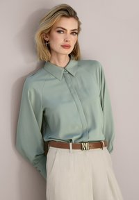 Hellgrüne Bluse mit Kragen, langen Ärmeln und subtiler Schimmer; kombiniert mit beigen Hosen und einem braunen Gürtel mit einer goldenen Logospange.