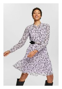Vestido con volantes en lavanda claro con patrón floral negro, mangas largas y cintura ajustada resaltada por un cinturón negro. Tejido suave y transparente.
