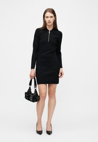 Γυναίκα με μαύρο μίνι φόρεμα μακρυμάνικο με φερμουάρ μπροστά, κρατώντας μαύρη τσάντα με τρούκς, στέκεται μπροστά σε απλό λευκό φόντο.