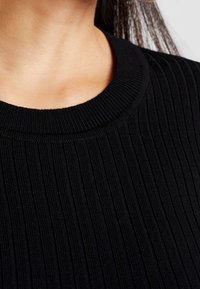 Gros plan sur une personne portant un pull en maille noire côtelée avec un col rond, les longs cheveux sombres partiellement visibles.