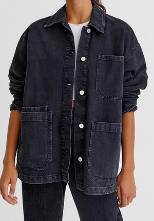 Femme portant une veste en jean noire oversize avec de grandes poches avant, des boutons argentés, des manches retroussées et un top court blanc en dessous.