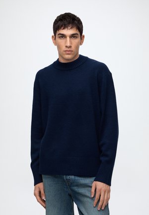 Homme aux cheveux courts et foncés portant un pull bleu marine et un jean bleu clair, debout devant un fond clair uni.