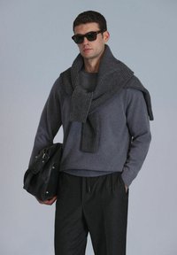 Grijze trui met een geribbeld sjaaltje eroverheen, gecombineerd met donkere broek. Het model houdt een zwarte tas vast en draagt donkere zonnebril.