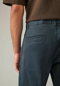Pantalones azul oscuro con un acabado texturizado, que presentan un bolsillo trasero y una cinturilla lisa, combinados con una manga de camisa marrón visible.