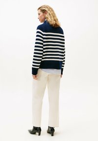 Frau mit schulterlangem blonden Haar, die einen navyblauen und weißen gestreiften Pullover, cremefarbene Hose und schwarze Stiefel mit Absatz trägt, steht und nach links schaut.