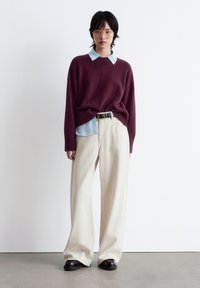 Ein bordeauxroter Strickpullover, der über einem hellblauen Hemd mit Kragen getragen wird, kombiniert mit cremefarbenen, weit geschnittenen Hosen und schwarzen Schuhen, vor einem schlichten Hintergrund.