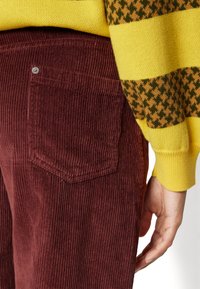Bordeaux corduroy broek met verticale ribbels, voorzien van een achterzak en een zilveren knop. Een geel en groen gestreept trui is gedeeltelijk zichtbaar.