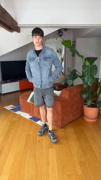 Ung mann kledd i denimbuksejakke og shorts står med hendene i lommene i en stue med tregulv og stor potteplante.