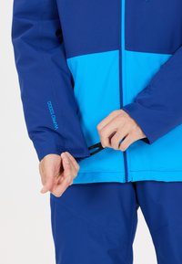 Blå och turkos vattentät jacka med dragkedja framtill, justerbara ärmslut och kardborreband. Slät textur och förstärkta söm detaljer.
