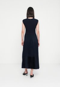 Kvinne med rett, mørkt hår iført en ermeløs svart topp, plissert marineblå midi-skjørt og svarte høyhelte slingback-sandaler, står med ryggen mot oss.