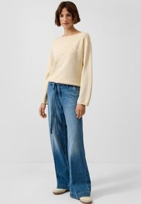 Frau mit kurzen braunen Haaren, die einen cremefarbenen Pullover mit Blattmustern, weit geschnittene blaue Jeans mit Gürtel und beige Schuhe trägt und auf einem weißen Boden steht.