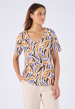 Femme portant un haut à manches courtes et col en V avec un motif abstrait de traits de pinceau orange, violet, marron et moutarde, associé à un pantalon beige clair.
