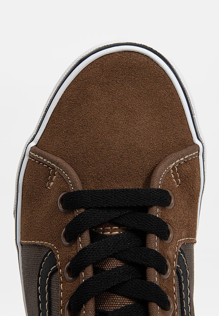 Zapatilla de ante marrón con cordones negros, suela blanca y panel lateral texturizado en contraste. Detalle de costura visible y forma de punta redonda.