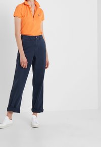 Orange pikétröja med krage och tre knappar, kombinerad med marinblå byxor, uppvikta vid manschetterna, och vita sneakers.