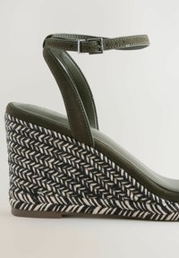 Sandalo con zeppa in velluto verde, con base in corda intrecciata nera e bianca. Presenta una cinghia alla caviglia con elementi in argento e una soletta liscia e imbottita.