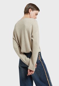 Maglione in pelliccia beige oversize con scollo ampio e polsini a coste, abbinato a jeans blu scuro con dettaglio a cucitura sfilacciata sul lato.