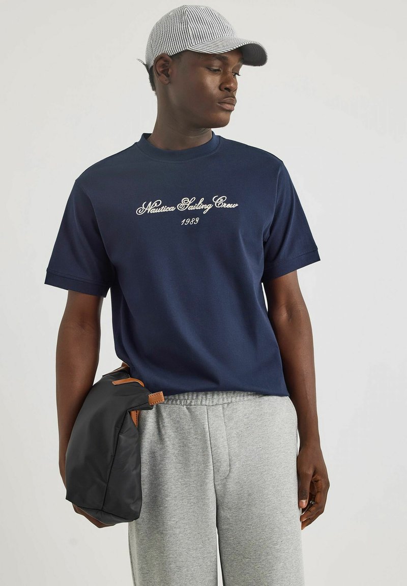 T-shirt in cotone blu marino con la scritta "Nautica Sailing Crew 1983" ricamata in bianco, abbinato a pantaloni della tuta grigi e a una borsa nera con dettagli marroni.