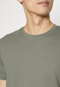 Detailní záběr osoby nosící texturované olivově zelené tričko s krátkými rukávy a kulatým výstřihem, zobrazující krk, čelist a horní část hrudníku.