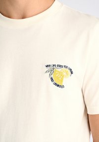 T-shirt color crema con maniche corte presenta una grafica di limone ricamata e testo sul lato sinistro del petto. Materiale in cotone morbido.