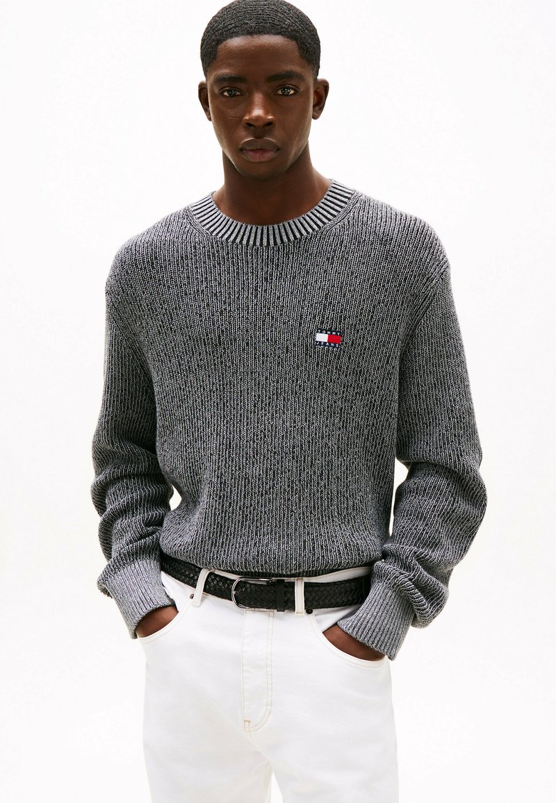 Maglione grigio a maglia con scollo e polsini a costine. Presenta un piccolo logo ricamato. Indossato con pantaloni bianchi e una cintura nera intrecciata.