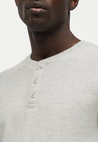 Κοντινό πλάνο ενός ατόμου που φοράει ανοιχτογκρι υφασμάτινη μπλουζά Henley με τρία ξεκουμπωμένα κουμπιά στον γιακά.