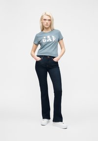 Νεαρή γυναίκα που στέκεται με τα χέρια στις τσέπες, φοράει μπλε μπλουζάκι "GAP", σκούρο μπλε τζιν και λευκά παπούτσια πλατφόρμα σε λευκό φόντο.