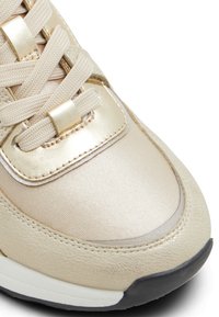 Zapatilla beige claro con acentos metálicos dorados. Presenta un upper sintético suave, áreas de cuero texturizado y cordones redondos. Suela de goma negra.
