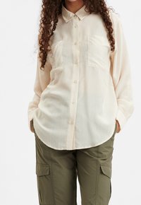 Camicia button-up crema in tessuto texture, con due tasche frontali e colletto classico. Abbinata a pantaloni cargo verde oliva.