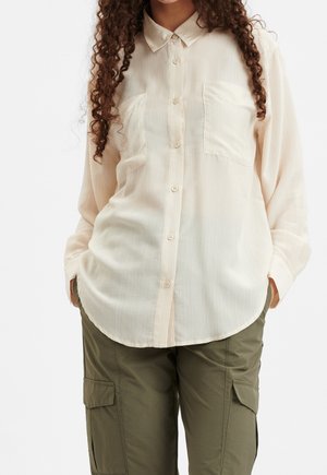 Chemise crème à boutons avec un tissu texturé, deux poches avant et un col classique. Assortie à un pantalon cargo vert olive.