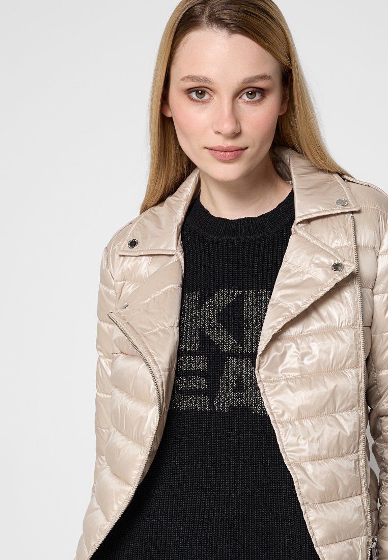 Helle beige, gepolsterte Jacke mit glänzendem Finish, Reißverschluss und Druckknöpfen. Getragen über einem schwarzen Strickpullover mit Textmuster.