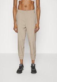 Beige, zulaufende Hose mit elastischen Bündchen, hergestellt aus einem leichten Stoff. Verfügt über einen Bund mit vorderen Falten. Kombiniert mit schwarzen Sportschuhen.